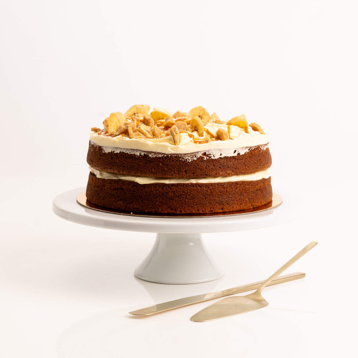 Side view of moist banana cake with cream cheese frosting and caramel drizzle by Sweet Talk, displayed on a cake stand, available for delivery in Auckland.