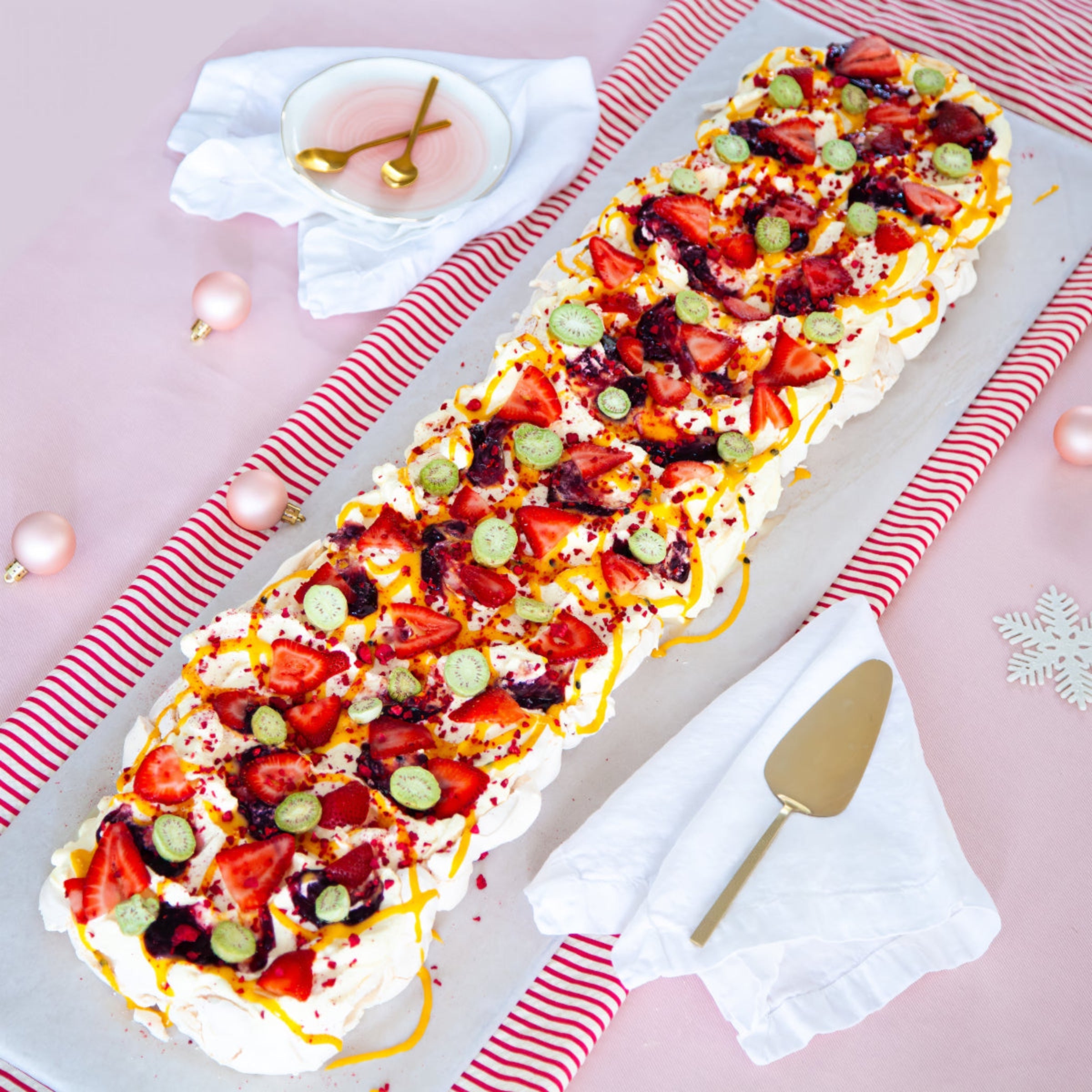 Long Christmas pavlova styled on a pink table with festive decorations, topped with cream, strawberries, kiwi berries and gold baubles.